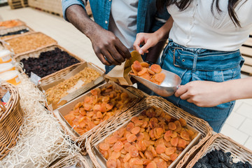cropped view of woman holding metal scoop with dried apricots near african american man with paper bag