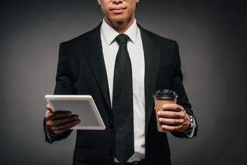 partial view of african american businessman holding coffee to go and digital tablet on dark background