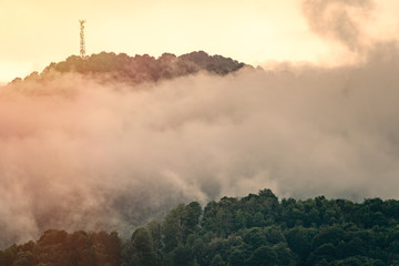 The green slope of the mountain with the cable cars disappeared in the fog. Orange sunset in the mountains.