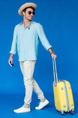 full length view of smiling traveler in safari hat and sunglasses holding yellow suitcase on blue
