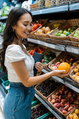 selective focus of cheerful asian woman looking at orange near fruits in store