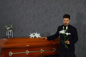 Sad young man with white lilies near casket in funeral home