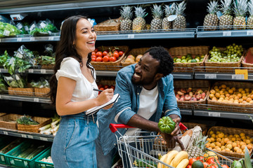 happy african american man looking at asian woman with notebook near shopping cart with groceries