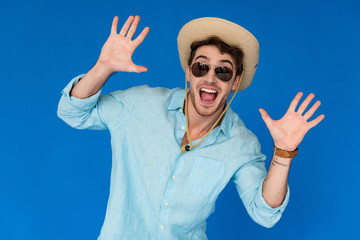 Naklejka premium front view of excited traveler in safari hat and sunglasses standing with hands up isolated on blue