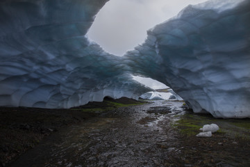 Small river flowing through the arch of blue snow. Ice bridge with big holes on the cloudy grey sky background. Landmannalaugar, Iceland