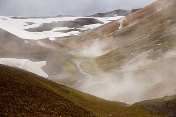 Small river flowing through the geyser valley with snow in Iceland. Beautiful landscape with steam column and dramatic sky background. Landmannalaugar, Iceland