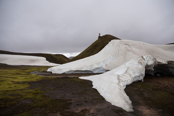 Small silhouette of a man on top of the mountain showing direction. Hills, green field, river and snow with cloudy background. Landmannalaugar trek, Iceland