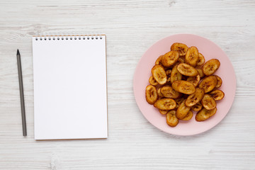 Fototapeta na wymiar View from above, homemade fried plantains on a pink plate, blank notepad on a white wooden surface. Flat lay, top view, from above. Copy space.