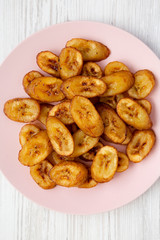 Overhead view, homemade fried plantains on a pink plate over white wooden background. Flat lay, overhead, from above. Close-up.