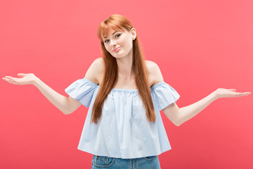 surprised redhead girl showing shrug gesture and looking at camera isolated on pink