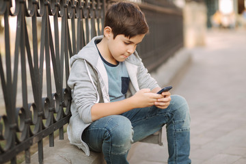 Young boy play online game at smartphone outside. Boy sitting in centre of the city. Boy play games while his mom going to the shop
