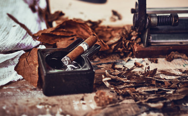 close up of a Cuban cigar and a black ceramic ashtray on the wooden table whit dried and cured tobacco leaves.