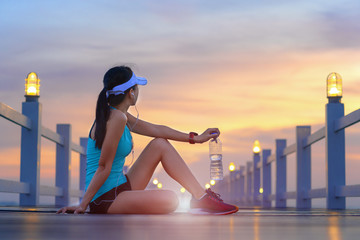 woman sport runner sitting on the wooden sea bridge holding drinking water in hand, resting or paused jogging for a while on the jetty, enjoy exercise jogging in sunset light