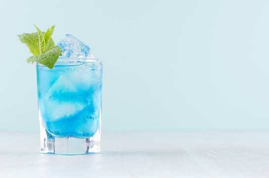 Exotic cold shot glass cocktail with blue curacao, ice cubes, green mint on soft light mint color background and white wood board.