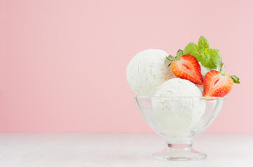 Three creamy ice cream balls in elegant glass bowl with green mint, strawberry pieces on white wood table and pastel pink wall.