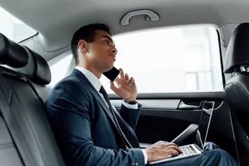 african american businessman in suit using laptop while talking on smartphone in car