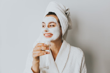 Face and body skin care, face treatment at home concept, wearing bathrobe and towel. Beautiful young woman drinking a fresh glass of water at home