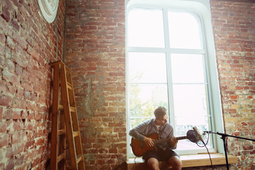 Fototapeta premium Young man recording music video blog, home lesson or song, playing guitar or making broadcast internet tutorial while sitting in loft workplace or at home. Concept of hobby, music, art and creation.