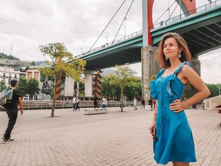 A young girl with long hair and a dress stands near the red bridge, the Museum and the square with a spider sculpture in Bilbao , Basque Country, Spain