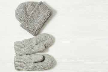 Knitted grey mittens and hat on white wood background. Warm clothing. Top view. Copy space.