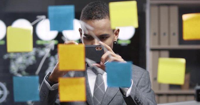 Young african man are standing in a modern office, look at the glass board with sticky notes and takes a picture on smartphone.