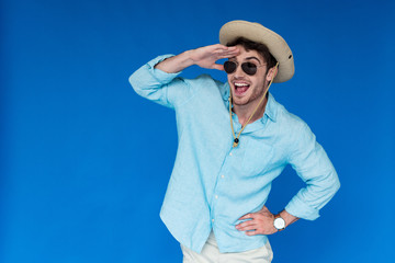 excited tourist in safari hat and sunglasses laughing and looking in distance isolated on blue