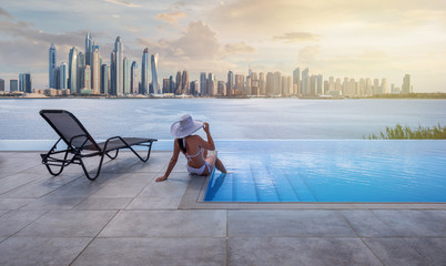 Beau panorama sur les toits de la marina de Dubaï en arrière-plan avec une piscine, une chaise longue et une femme avec un chapeau blanc au coucher du soleil.