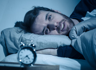 Young man in bed staring at alarm clock trying to sleep feeling stressed and sleepless