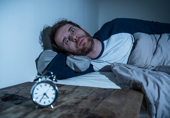 Desperate stressed young man whit insomnia lying in bed staring at alarm clock trying to sleep