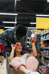 selective focus of asian girl with duck face touching sunglasses and holding bottle with wine while sitting in shopping cart near african american man