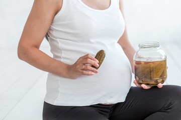 Pregnant woman is holding salted cucumbers in a jar near her belly.