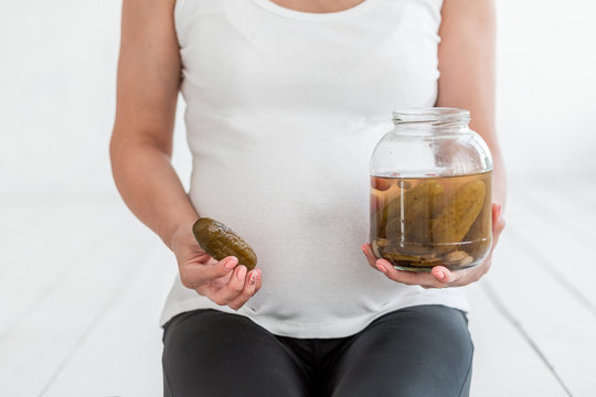 Pregnant woman is holding salted cucumbers in a jar near her belly.