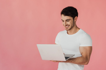 smiling muscular man in white t-shirt using laptop isolated on pink