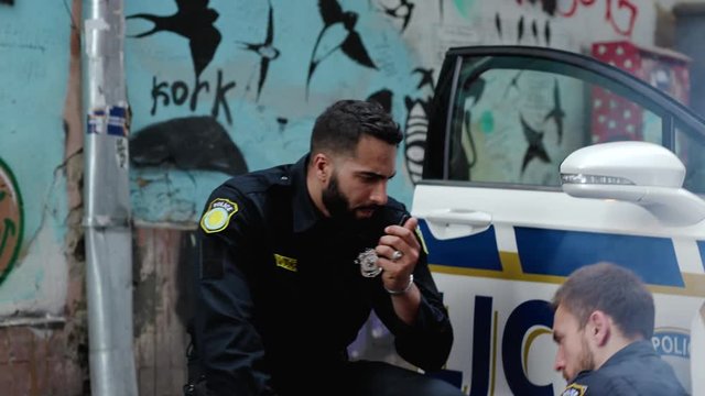 Dangerous city. View of policeman injured in shootout leaning on the car crying of pain. His friend cop talks walkie-talkie call for help protect his colleague.