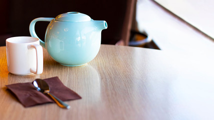 White empty cup and blue tea pot, and defocused spoon on brown paper napkin on wooden table in restaraunt. Selective focus. Horizontal banner with copy space.