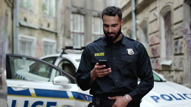 Portrait of joyful sociable young policeman browsing on smartphone in the street. Police cop with mobile phone against a patrol car.