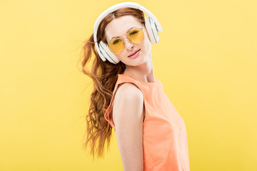 attractive redhead woman in sunglasses listening music in headphones isolated on yellow