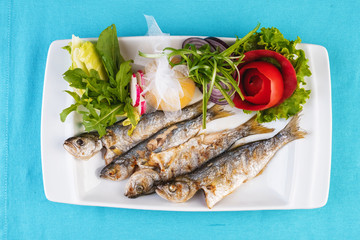 Mediterranean dish, European cuisine. Whole fish grilled capelin, served with a salad of vegetables — greens, arugula, onion rings, tomato, green and yellow pepper and lemon