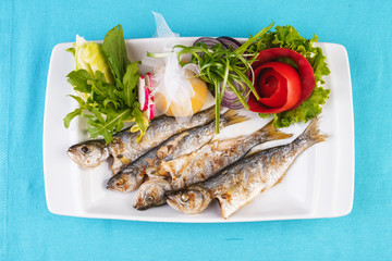 Mediterranean dish, European cuisine. Whole fish grilled capelin, served with a salad of vegetables — greens, arugula, onion rings, tomato, green and yellow pepper and lemon