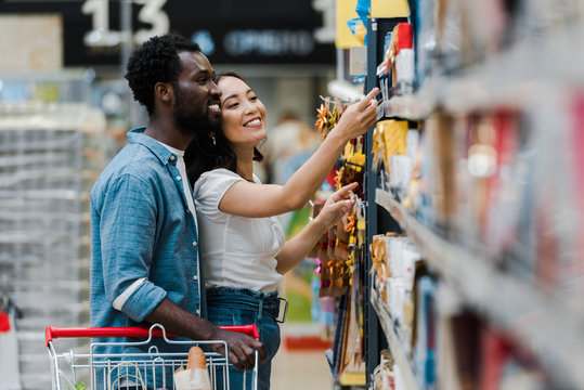 Selective focus of happy African American man standing with Asian woman pointing with finger at groceries in supermarket