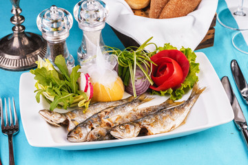 Mediterranean dish, European cuisine. Whole fish grilled capelin, served with a salad of vegetables — greens, arugula, onion rings, tomato, green and yellow pepper and lemon