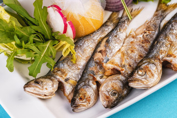 Mediterranean dish, European cuisine. Whole fish grilled capelin, served with a salad of vegetables — greens, arugula, onion rings, tomato, green and yellow pepper and lemon