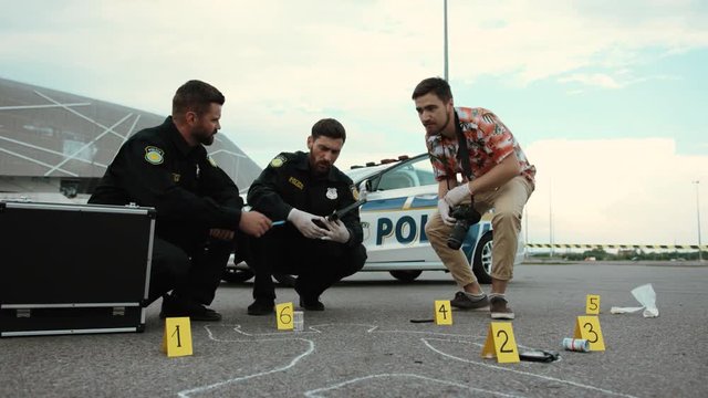 Low angle view of handsome men at work. Two policemen and CSI investigator collect and examine evidence near the body chalk outline on the ground.