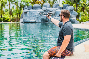 Bearded man launches drone for flight, with which you can take photos and video filming, near the swimming pool and palm trees, side view.