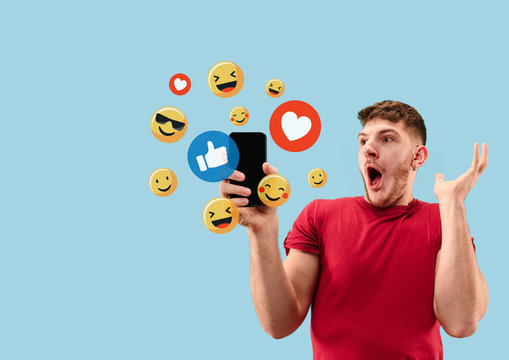 Social media interactions on mobile phone. Internet digital marketing, Chating, commenting, liking. Smiles and icons above smartphone screen, that holding by young man on blue studio background.