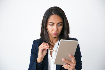 Fototapeta premium Focused pensive business woman studying report on tablet. Beautiful Latin woman in office jacket holding gasses and looking at tablet screen. Digital technology concept