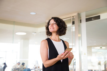 Cheerful woman waiting someone in hotel lobby. Happy middle aged woman standing in hall, consulting phone and staring into distance. Waiting concept