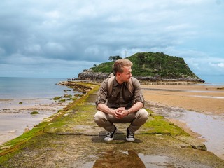 Blond man sitting on a stone overlooking the coastline of the Bay of Biscay, mountain view, beach, stones