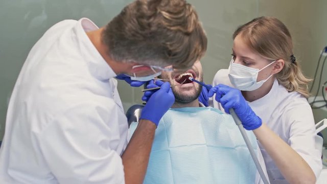 Attractive brunette man sitting in chair while having dental procedure with dentist and assistant at the dental clinic