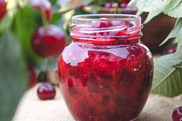 Cherry jam in a glass jar on a background of green leaves on a table of wooden boards.